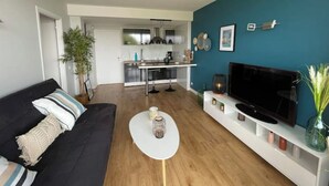 Una sala de estar moderna con un sofá negro, una mesa de centro blanca y un televisor sobre un mueble blanco. Se ve una cocina al fondo.