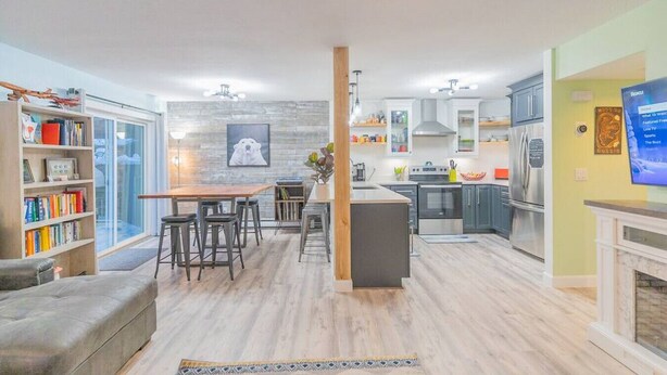 Una cocina moderna con una isla central, un comedor con mesa de madera y taburetes de bar, y una sala de estar con sofá y estantería.