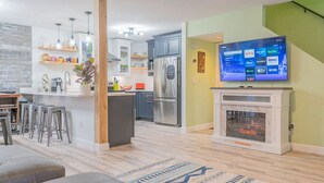 Una cocina moderna con electrodomésticos de acero inoxidable, una zona de bar con taburetes y una sala de estar con chimenea y televisor de pantalla plana que muestra servicios de transmisión.