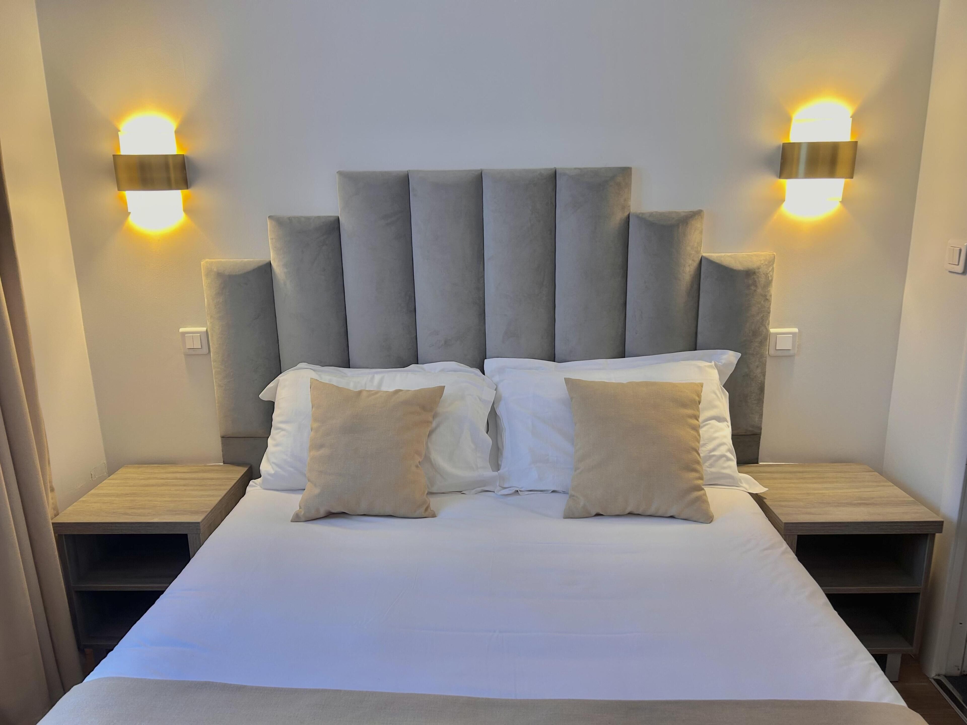 A bed with a grey upholstered headboard, two bedside tables, and wall-mounted lighting.