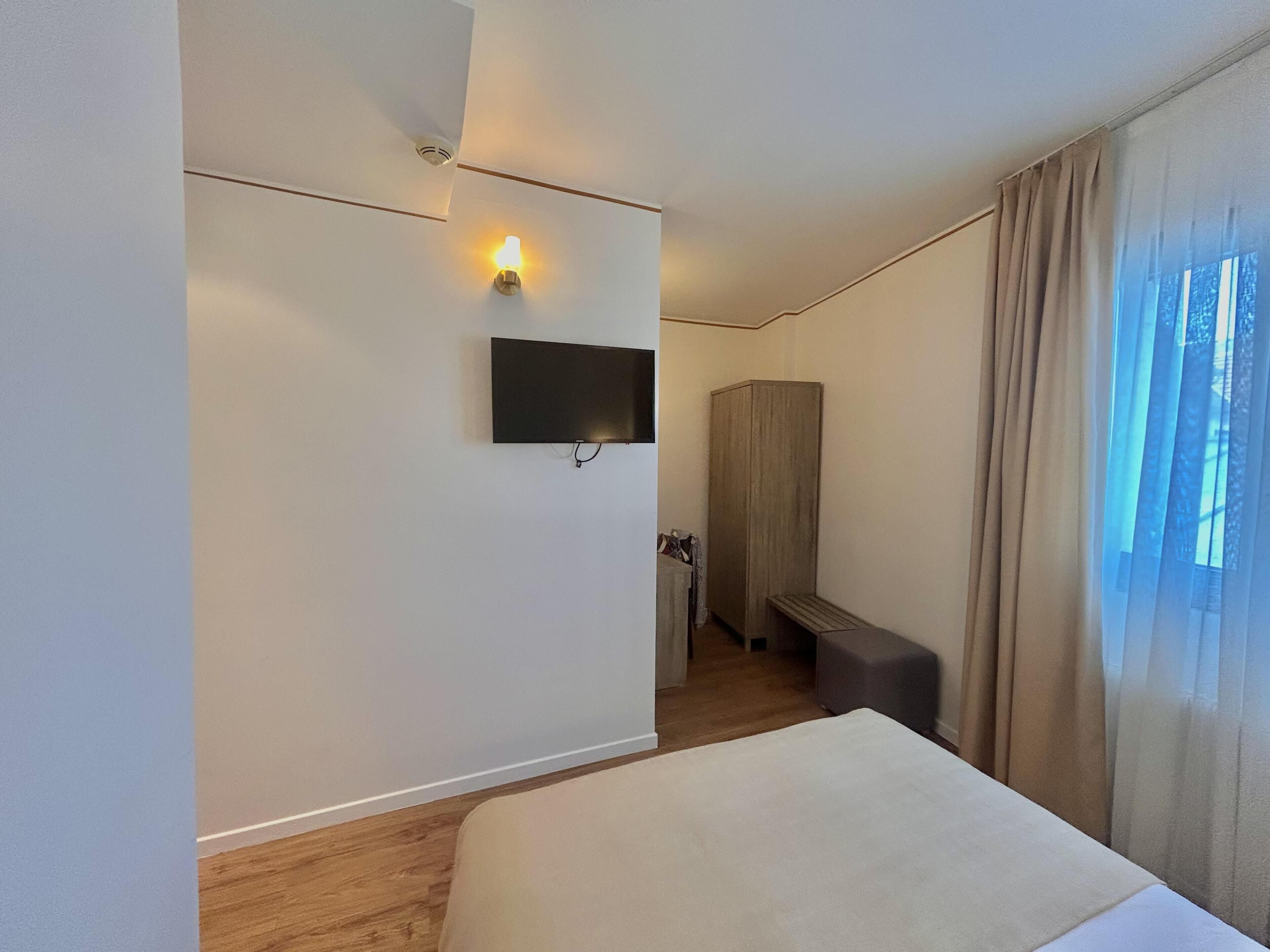 Habitación de hotel con cama, televisor montado en la pared, armario y ventana con cortinas.