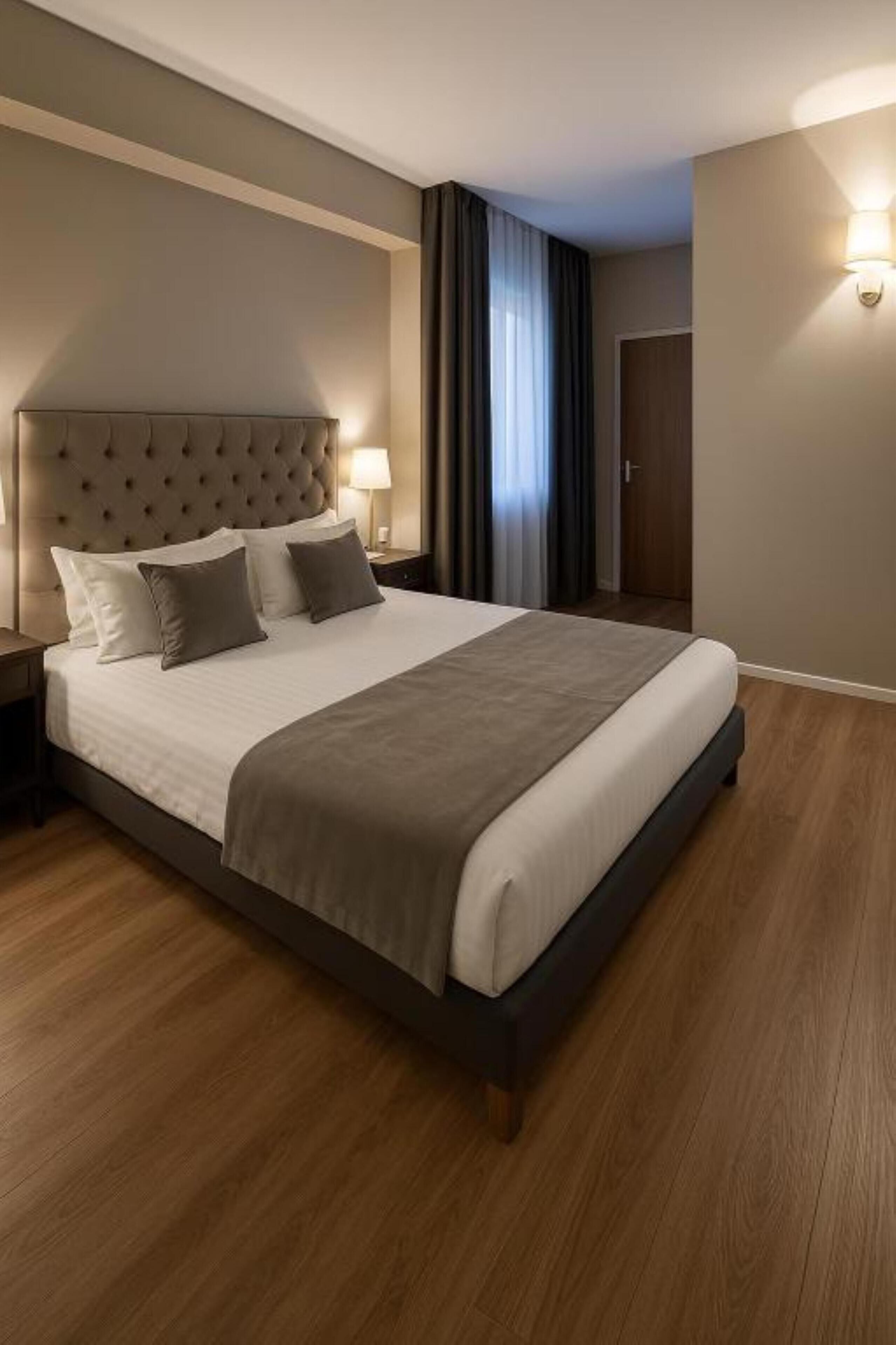Una cama grande con cabecera capitonada, una mesita de noche, una lámpara y vistas de una puerta y cortinas.