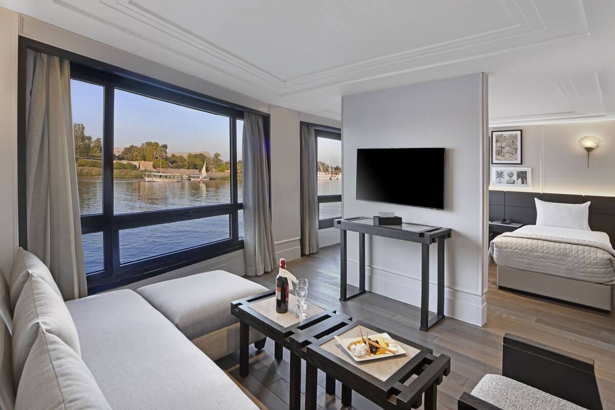 Une chambre d’hôtel moderne dotée d’une grande fenêtre donnant sur l’eau et offrant une vue sur les bateaux, d’un téléviseur à écran plat, d’un lit, d’un canapé et d’une petite table à manger avec une bouteille et des verres.