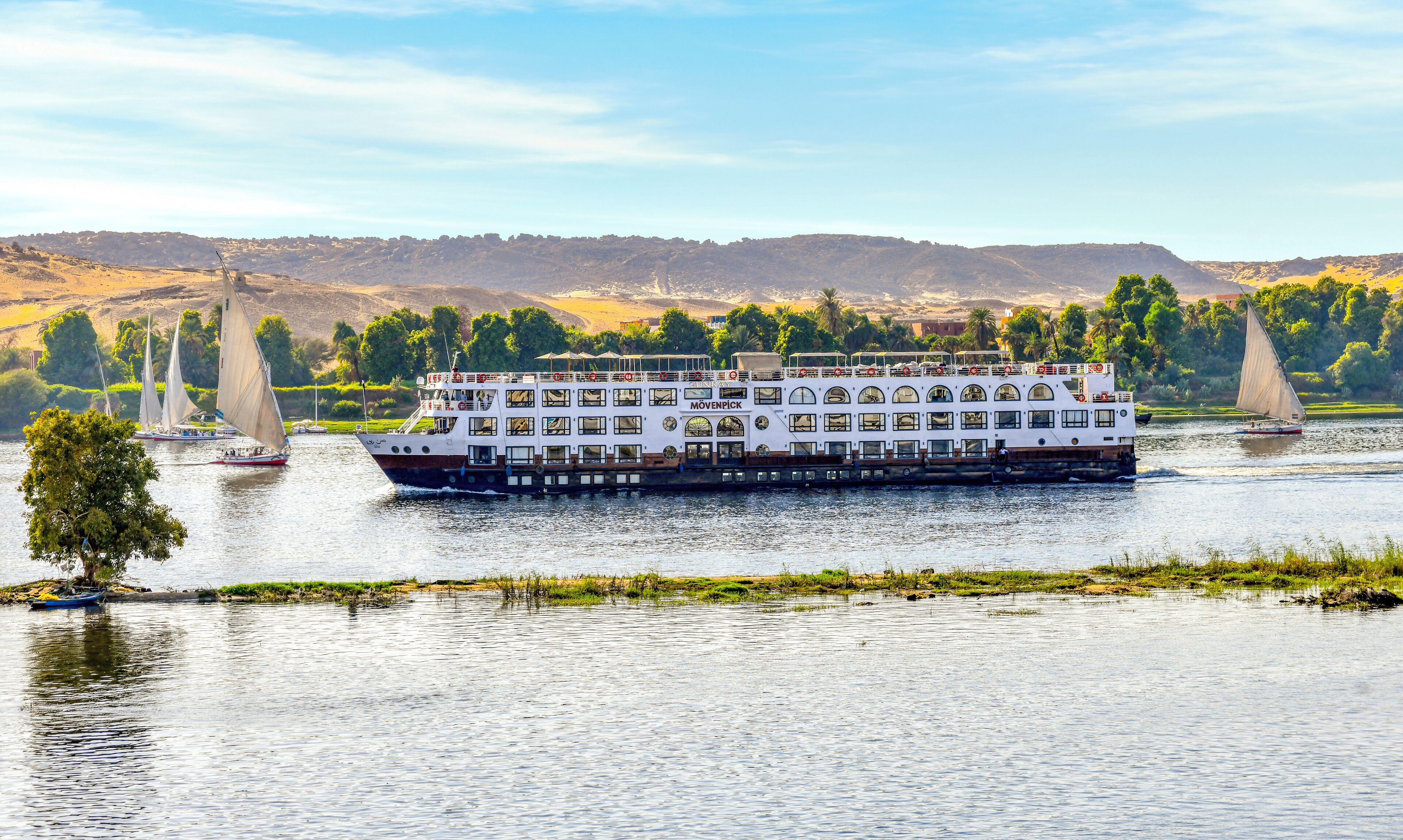 Un gran barco de crucero fluvial con varias cubiertas y un comedor, rodeado de agua tranquila y vegetación.