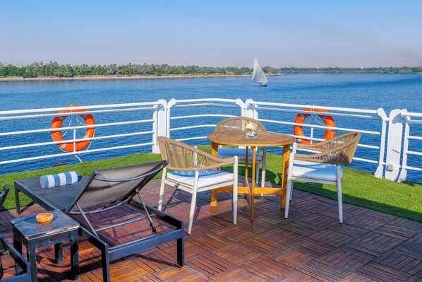 Una terrazza con posti a sedere all'aperto, inclusi lettini e un tavolo, con vista su un'area d'acqua e una vela all'orizzonte.
