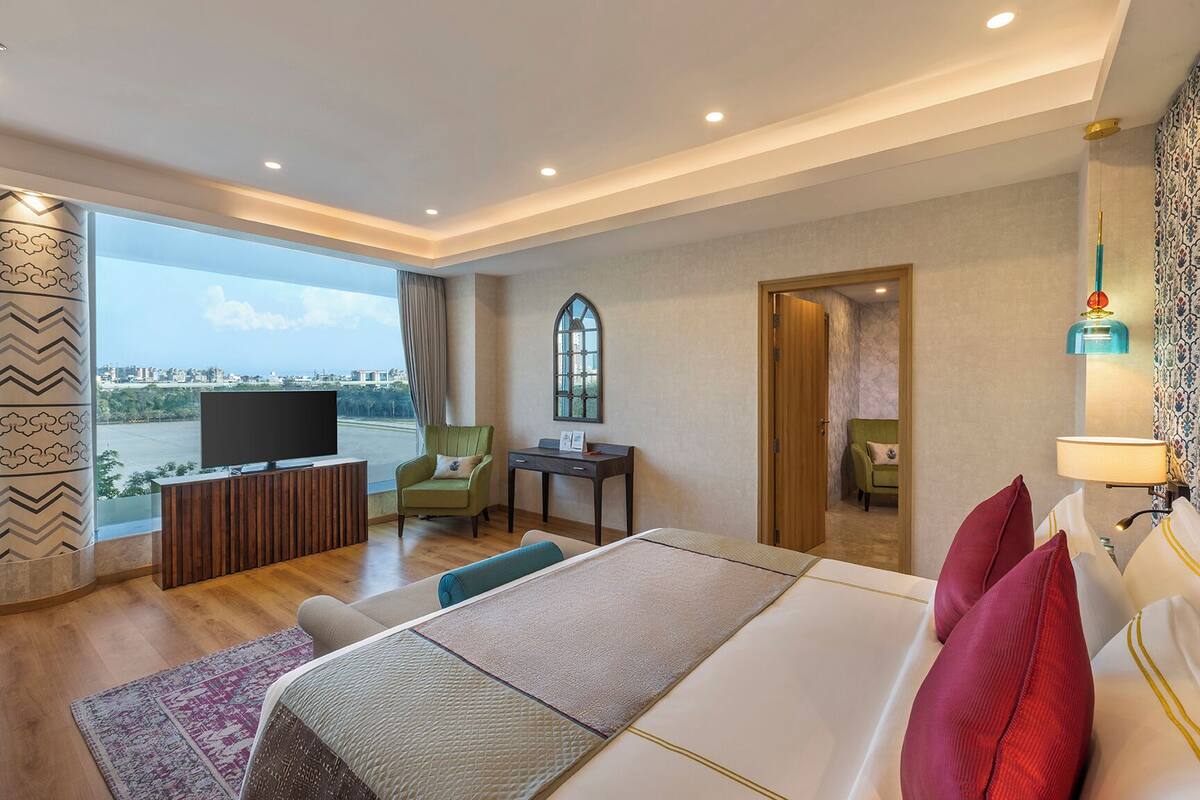 A hotel room with a large bed, a TV, a desk, a chair, and a view of the city.