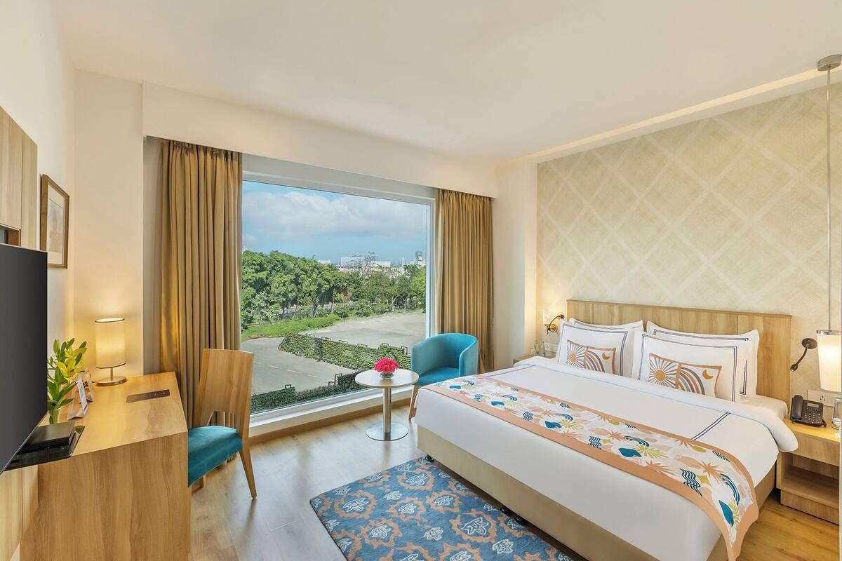 A hotel room with a bed, a desk, a chair, a small table with a vase, and a view of greenery and buildings.