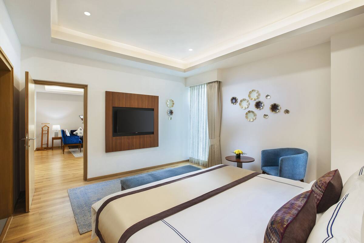 A modern hotel room with a large bed, a flat-screen TV, a blue armchair, and a small table with a vase of flowers.
