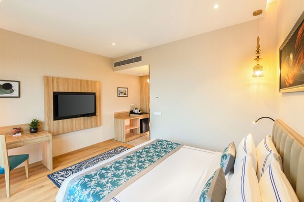 A modern hotel room with a flat-screen TV, a desk with a chair, a bed with a patterned bedspread, and a wall-mounted light fixture.