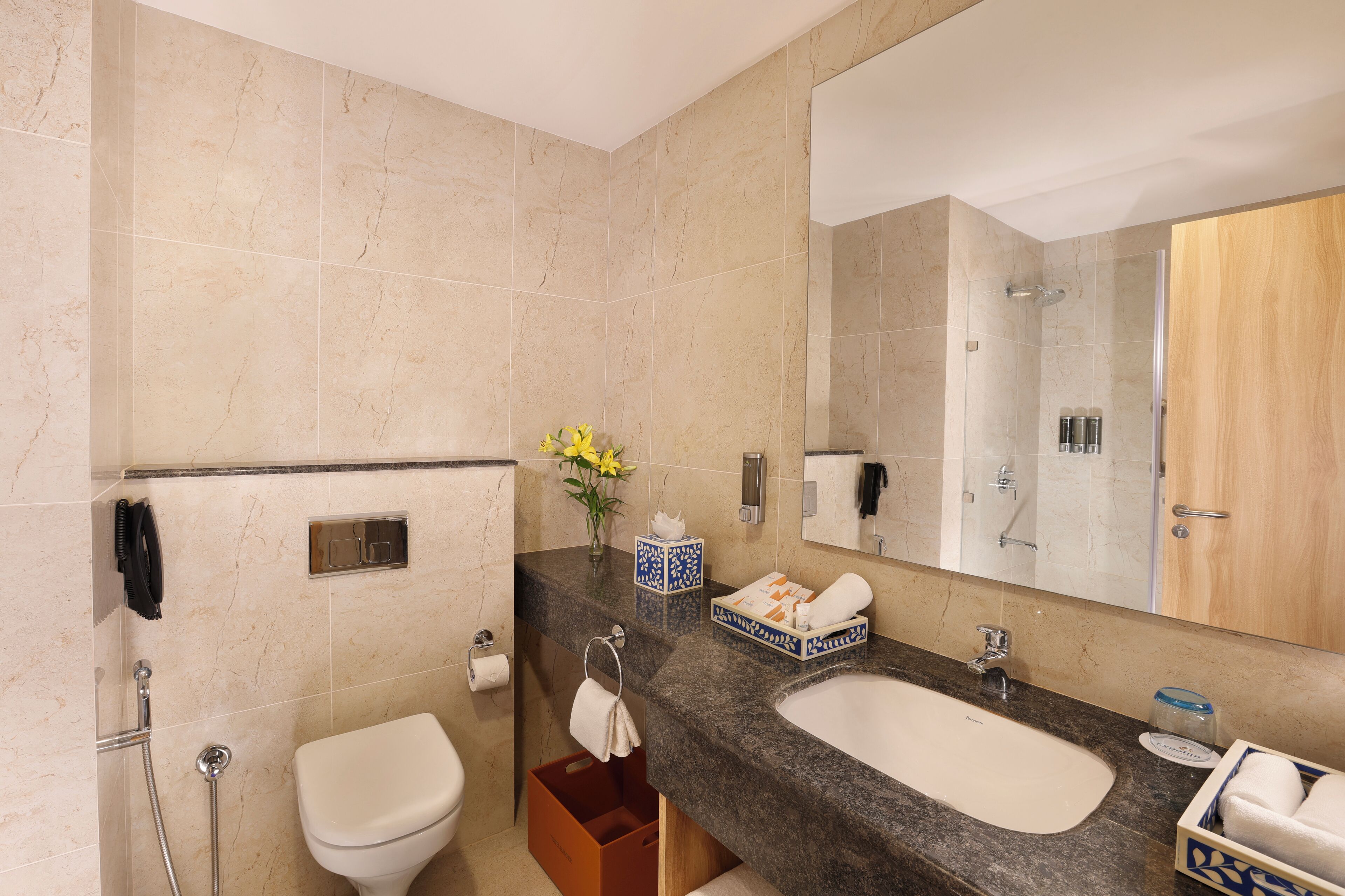 Een badkamer met een marmeren vloer, een wit toilet, een douche, een wastafel met spiegel en een vaas met gele bloemen.