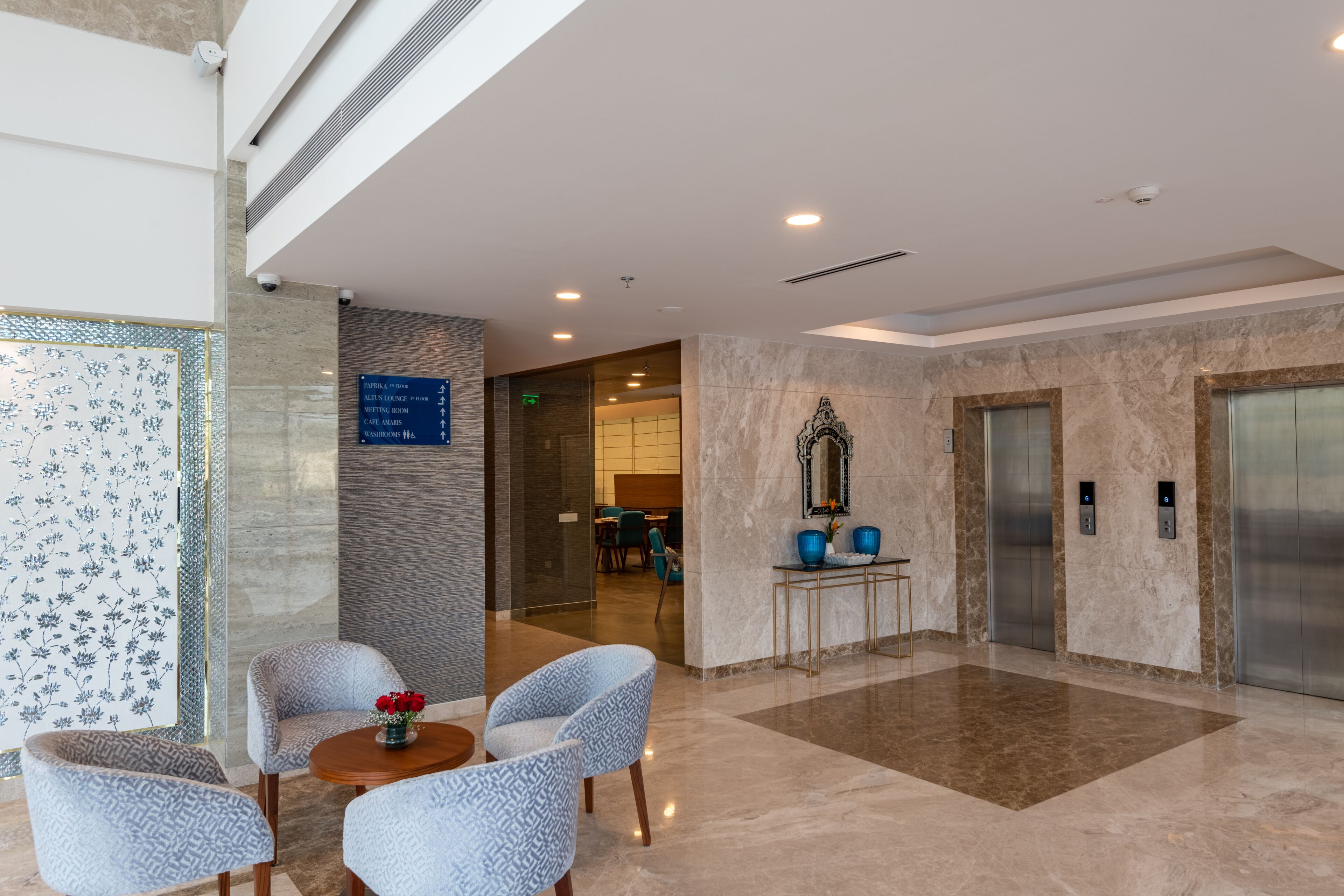 A modern lobby with a marble floor, patterned chairs, a small table with flowers, and an elevator.