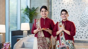 Two individuals in traditional attire holding a tray with offerings, standing in a modern room with a decorative wall and a sofa. - ExpoInn Suites and Convention (Greater Noida)