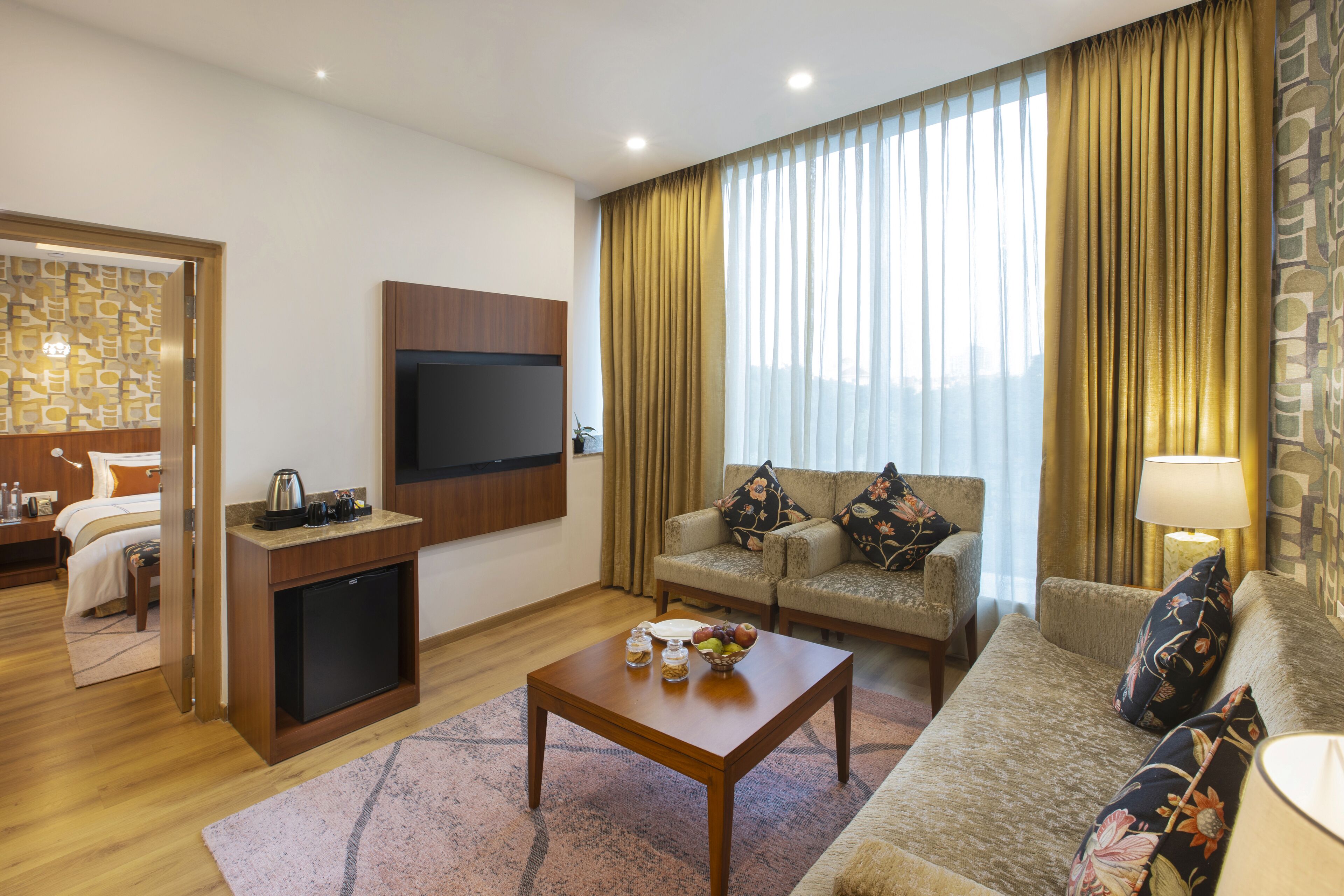 A hotel room with a wooden coffee table, a sofa, a TV mounted on the wall, and a small kitchenette.