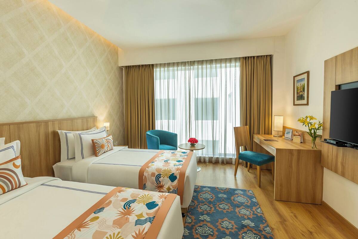 A hotel room with two beds, a desk, a chair, a TV, and a window with curtains.