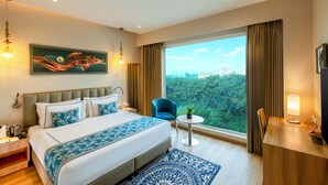 A hotel room with a large bed, a desk, a chair, a small table with a plant, and a view of a forest through a large window. - ExpoInn Suites and Convention (Greater Noida)