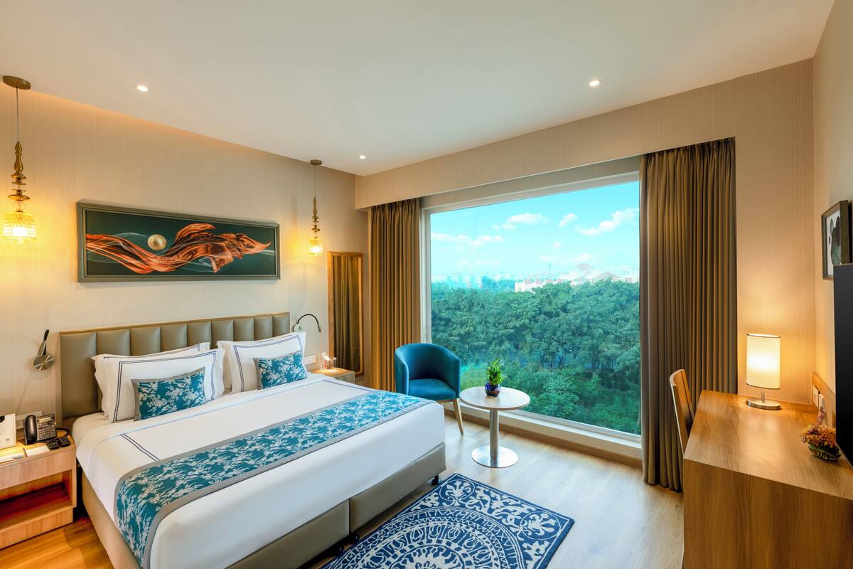 A hotel room with a large bed, a desk, a chair, a small table with a plant, and a view of a forest through a large window.