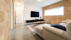 A modern living room with a sofa, a coffee table, a flat-screen TV mounted on a brick wall, and a large window providing natural light. - Whales & Wombats (Bass Coast)