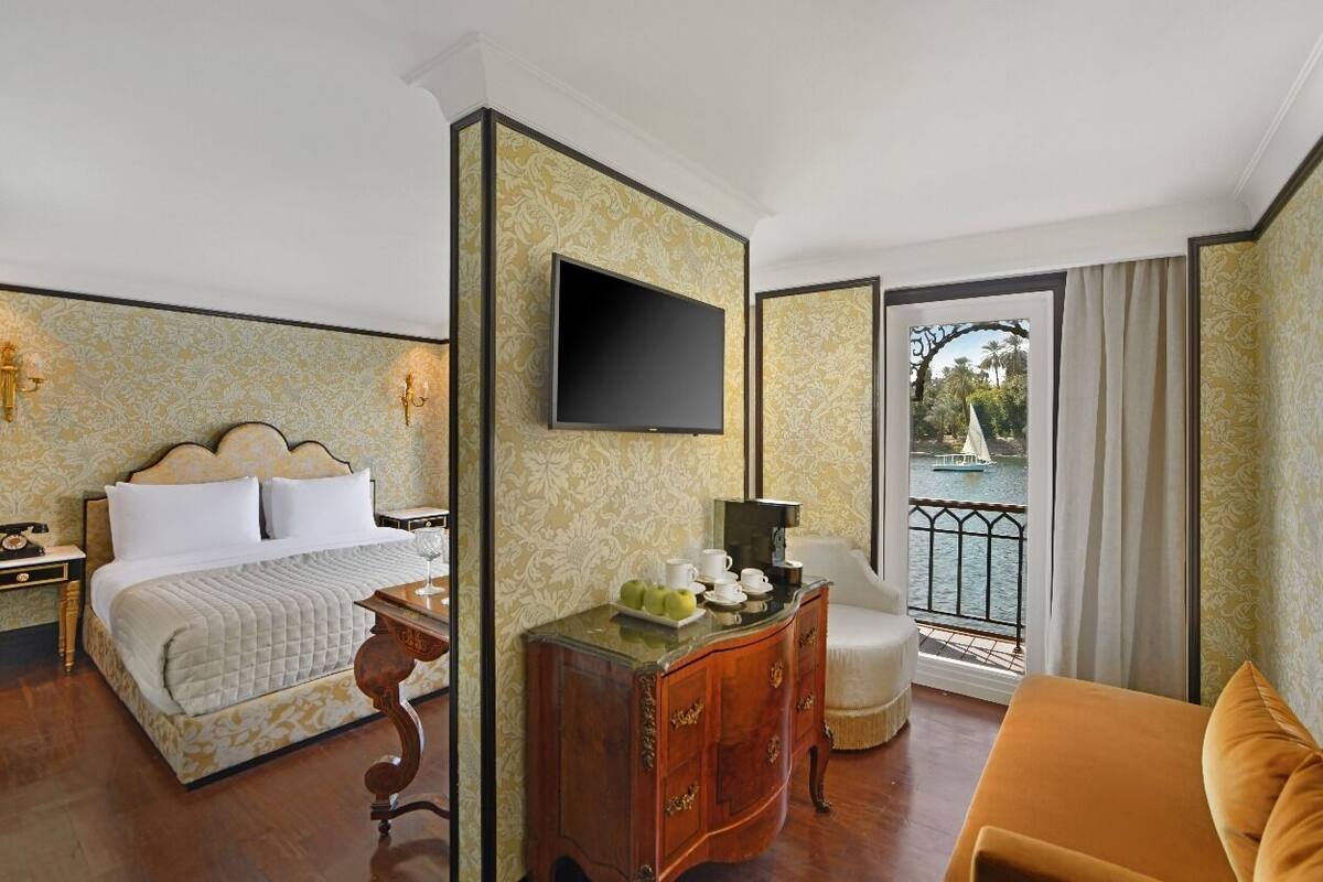 A hotel room with a bed, a desk with a flat-screen TV, a dresser, a chair, and a view of a body of water through a balcony.
