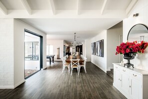 A spacious dining area with a long table and white chairs, leading to a kitchen area with a white cabinet and a vase of red flowers.