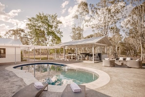Ein Swimmingpool mit Liegestühlen, einem überdachten Sitzbereich und Bäumen im Hintergrund.