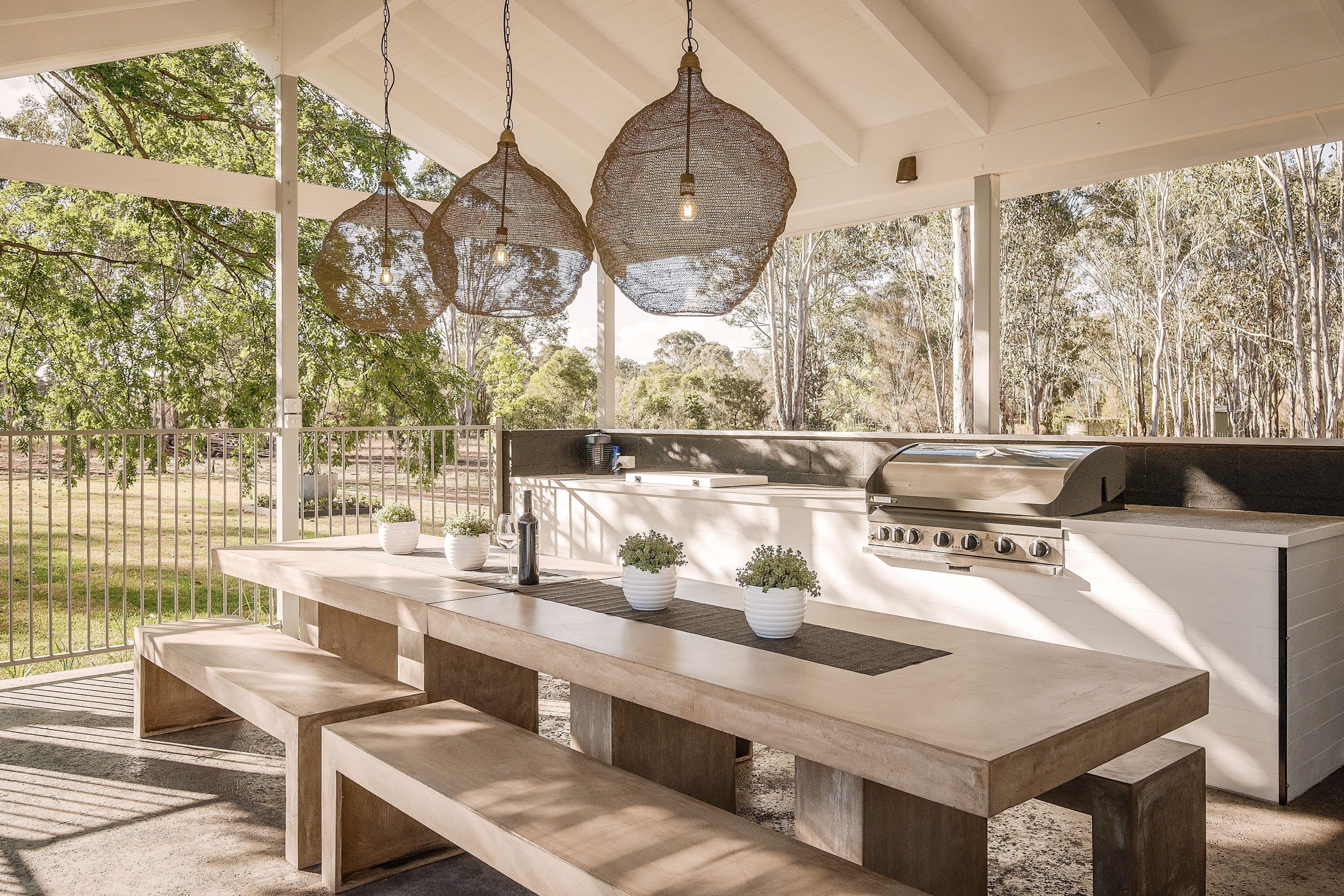 An outdoor kitchen area with a built-in grill, a long table with benches, hanging pendant lights, and potted plants.