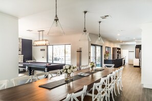 A spacious dining area with a long wooden table, white chairs, and pendant lighting.