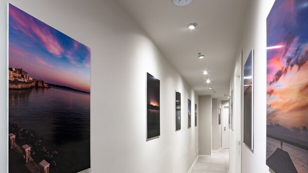 A hallway with a series of framed artworks on the walls, a white floor, and recessed lighting. - PhotoRooms B&B (Syracuse)
