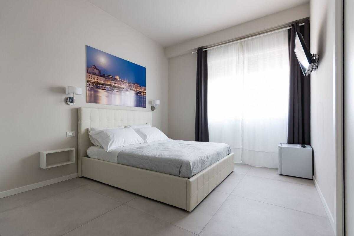 A modern hotel room with a large bed, a night view painting, a bedside table, and a window with curtains.