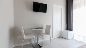 A modern room with a flat-screen TV mounted on the wall, a small white table with two chairs, and a window with curtains. - PhotoRooms B&B (Syracuse)