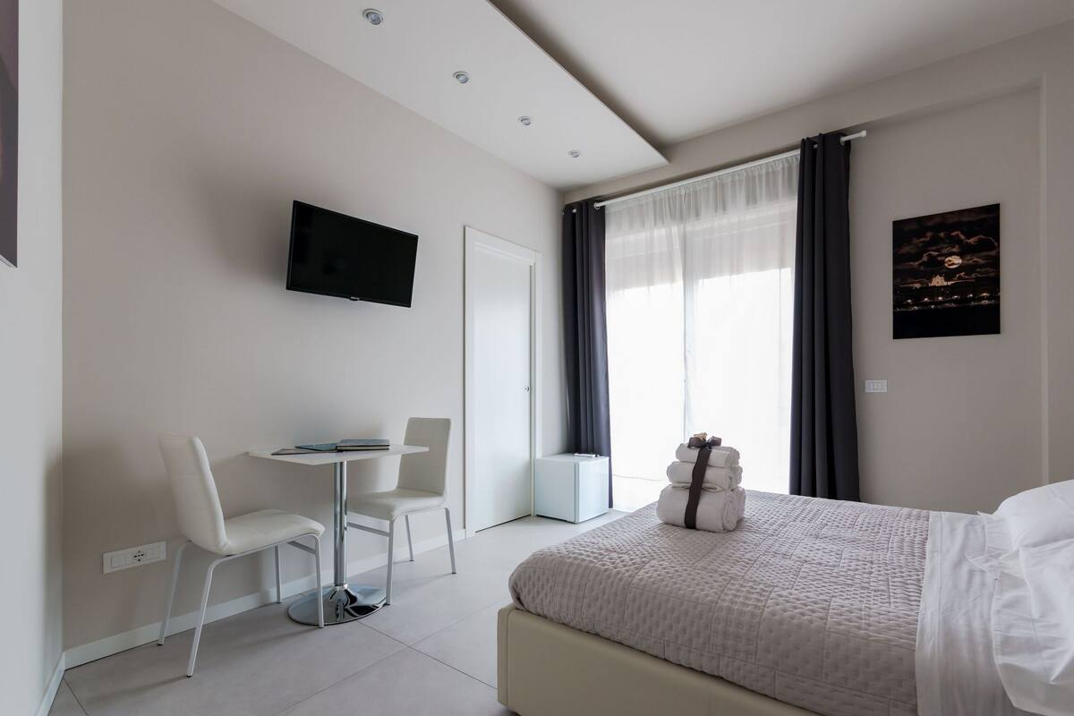 A modern hotel room with a bed, a desk with a chair, a television on the wall, and a window with curtains.