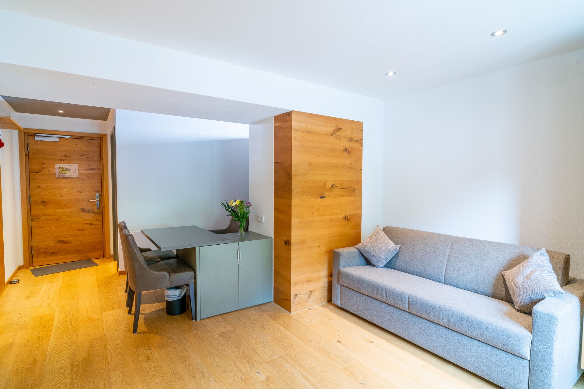 Een moderne woonkamer met een grijze bank, houten details en een eethoek met een tafel en stoelen.