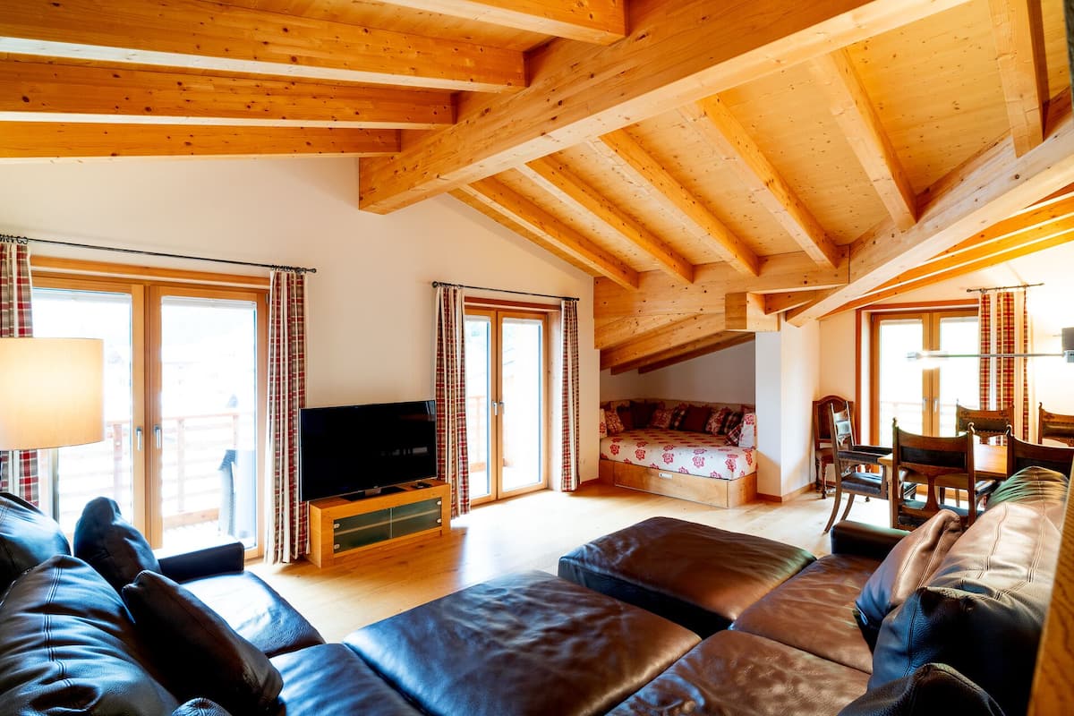 A spacious living room with a wooden ceiling, a sofa, a TV, and a bed in the background.