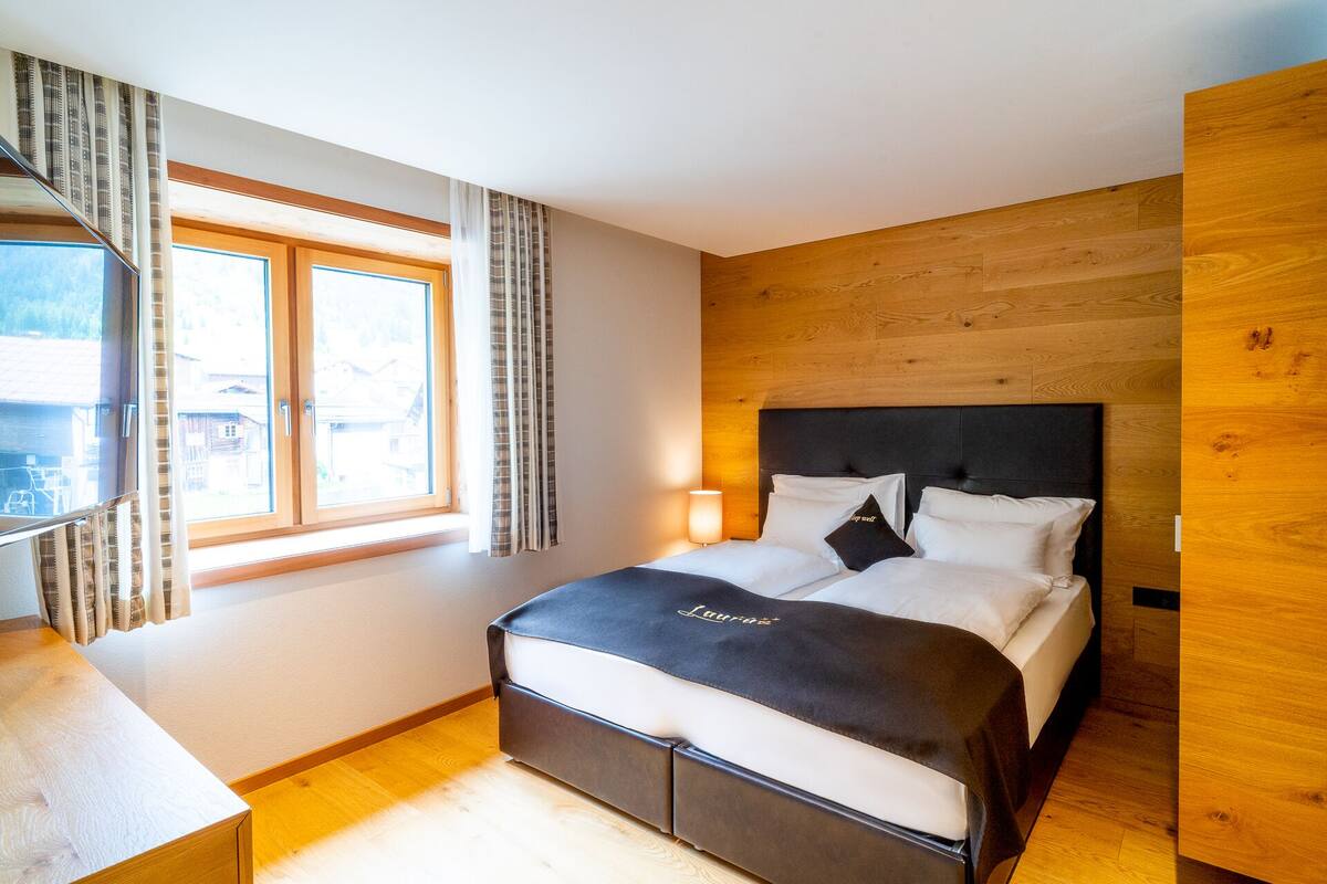 A modern hotel room with a large bed, wooden paneling, a window with curtains, and a bedside lamp.