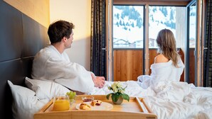 A couple in a hotel room with a wooden headboard, a large window with a view of snow-covered mountains, and a breakfast tray on the bed. - Apart-Hotel Laurus (Lech am Arlberg)