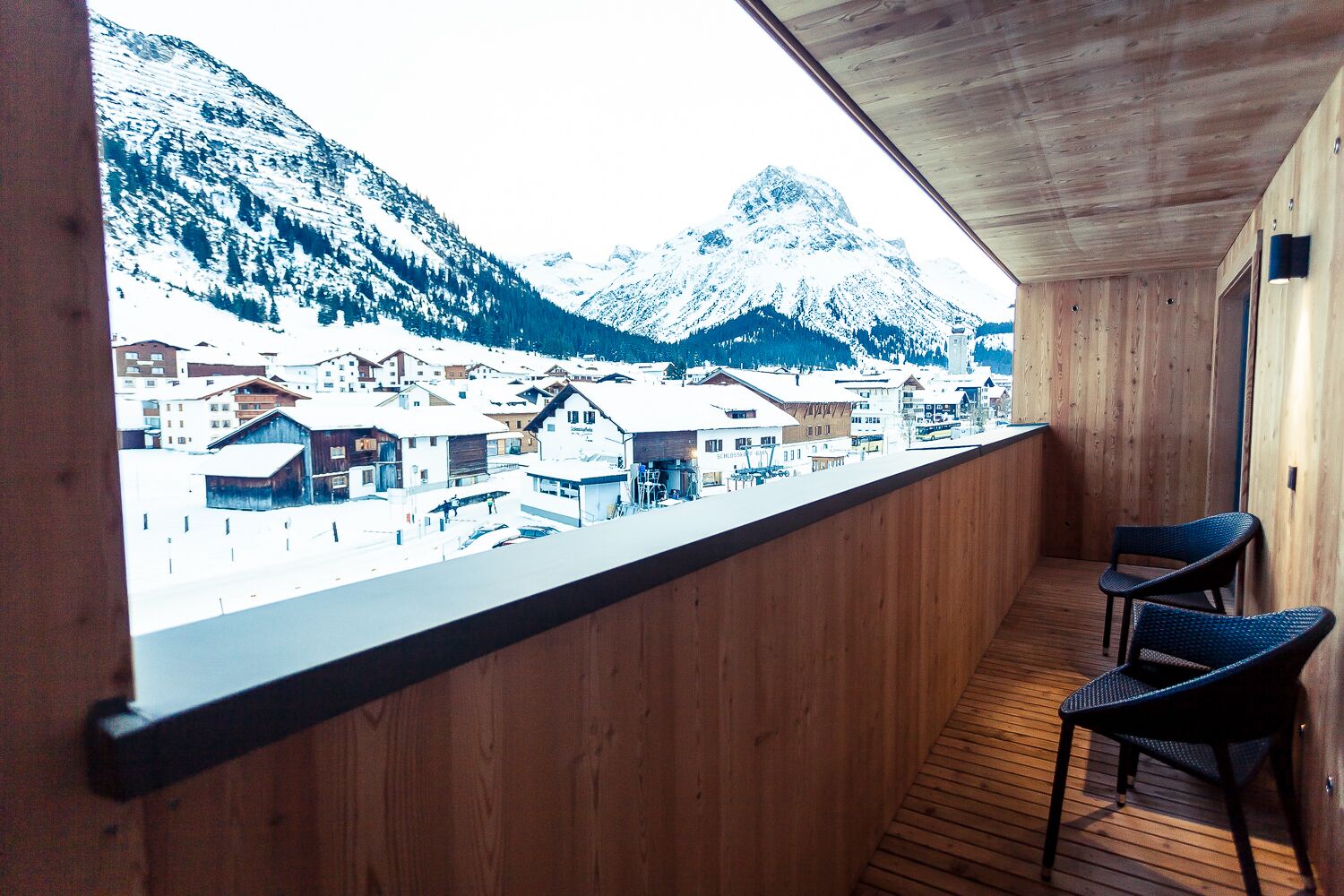 A balcony with wooden railings and two wicker chairs, overlooking a snowy mountain and a town.