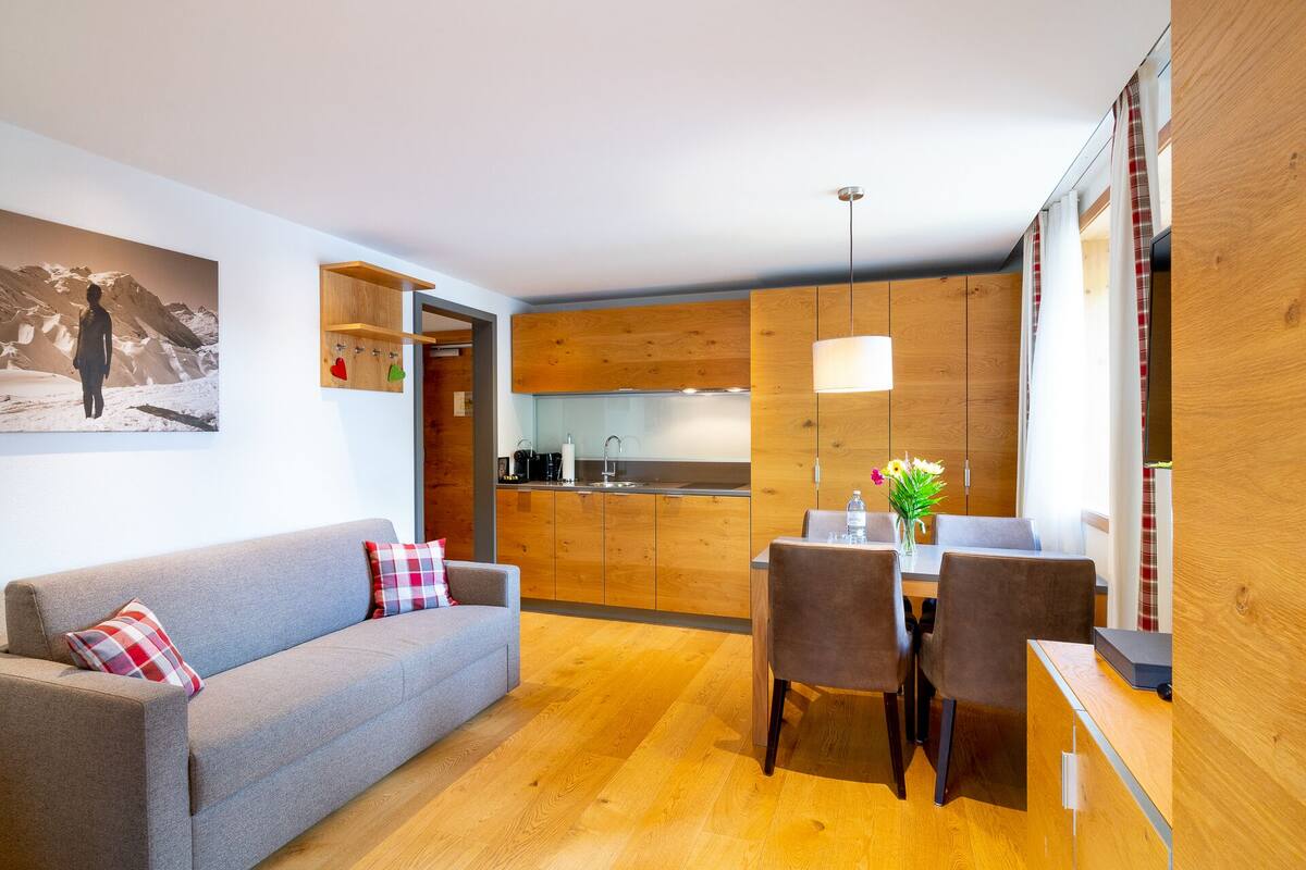 A modern living room with a grey sofa, a dining table with chairs, wooden cabinets, and a kitchen area with a sink and stove.