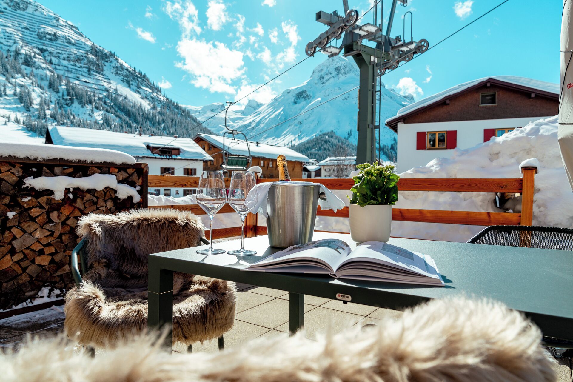 A table with two wine glasses, a metal bucket, and an open book, set against a snowy mountain backdrop with a ski lift and buildings.