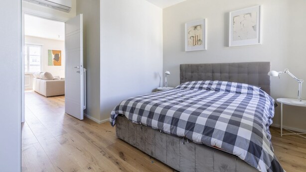 Un dormitorio con una cama grande, una mesita de noche y una silla. Se ve una puerta que da a otra habitación.
