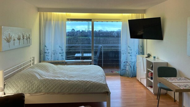A bedroom with a bed, a desk, a chair, a TV, and a balcony with curtains. - Apartment Thermen-zeit (Bad Fuessing)