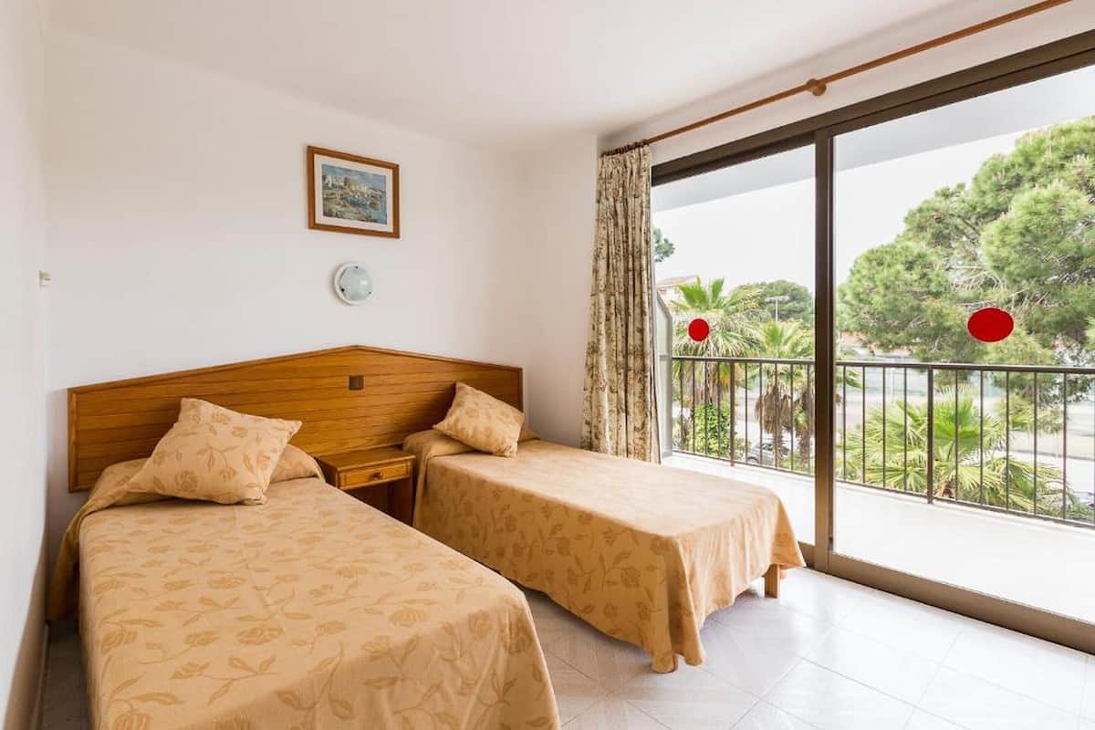 A hotel room with two beds, a wooden headboard, a small bedside table, a wall-mounted light, and a balcony with a view of trees and a railing.
