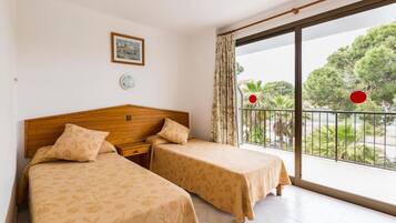 A hotel room with two beds, a wooden headboard, a small bedside table, a wall-mounted light, and a balcony with a view of trees and a railing.