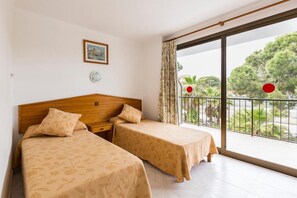 Una habitació d'hotel amb dos llits, un panell de fusta al capçal, una petita taula de nit, una làmpada penjada a la paret i un balcó amb vista a arbres i una barana.