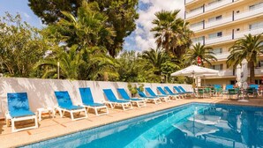 A swimming pool with lounge chairs and umbrellas, surrounded by palm trees and a building with balconies. - Bella Maria Apartments & Pool Club (Felanitx)