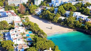 An aerial view of a coastal area with buildings, a beach, and clear turquoise waters. - Bella Maria Apartments & Pool Club (Felanitx)