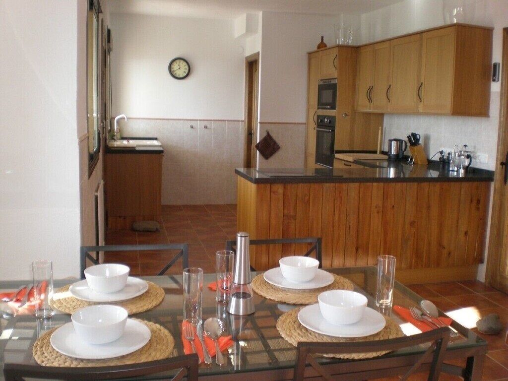 A dining area with a glass table set for four, surrounded by wooden chairs. A kitchen with wooden cabinets and a stove is visible in the background.