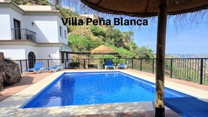 Una piscina con sillas de descanso, una sombrilla de paja y vistas a una colina.
