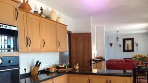 Una cocina con armarios de madera, encimera negra y estufa. Hay una zona de comedor con un sofá rojo y una mesa con flores.