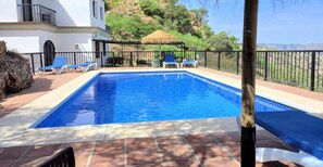 A swimming pool with lounge chairs, a thatched umbrella, and a view of a hillside.