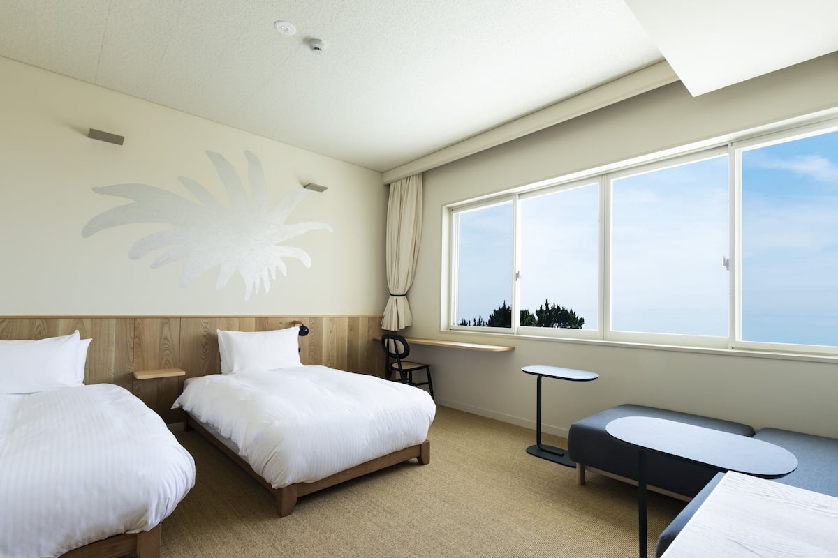 Habitación de hotel con dos camas, una mesita, un banco y un ventanal con vistas al cielo y a los árboles.