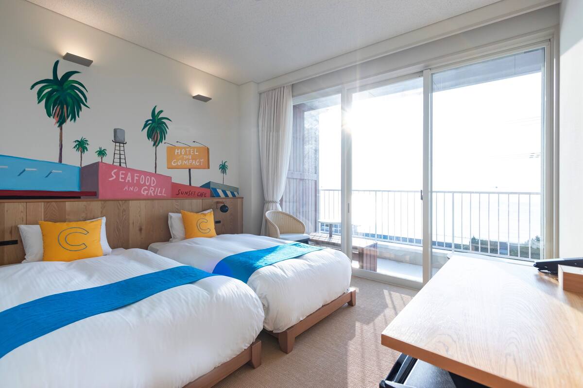 Habitación de hotel con dos camas, un escritorio y un balcón con vistas.